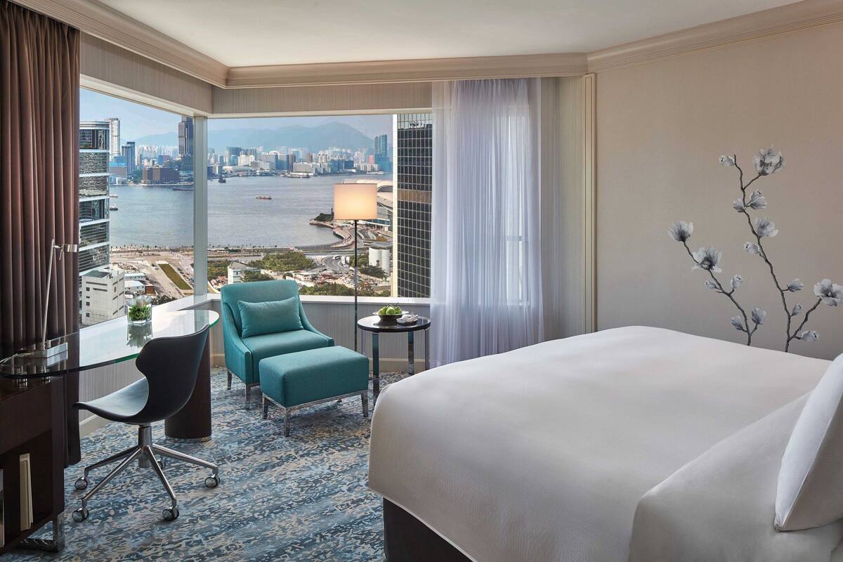 A hotel room with a large bed, a desk with a chair, a small table with a lamp, and a view of a cityscape and water.