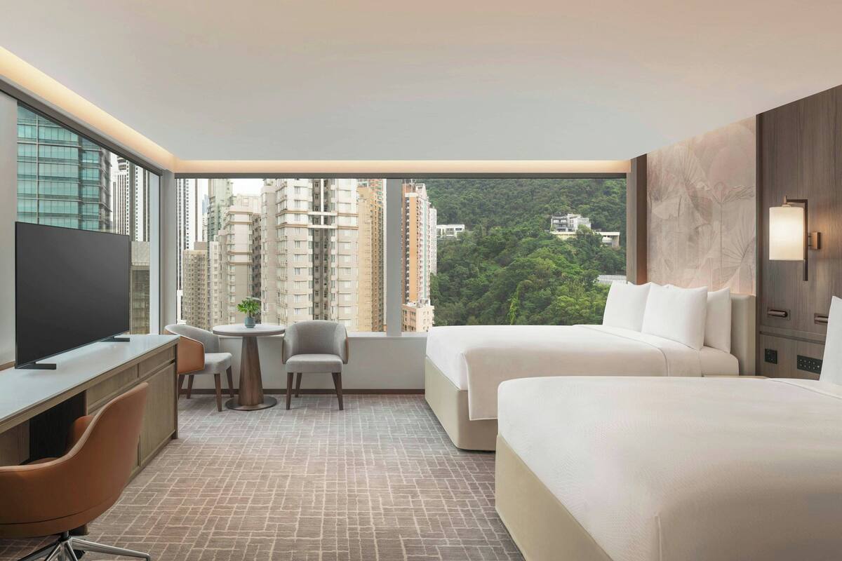 A hotel room with a large window, a desk with a chair, two beds, and a TV.