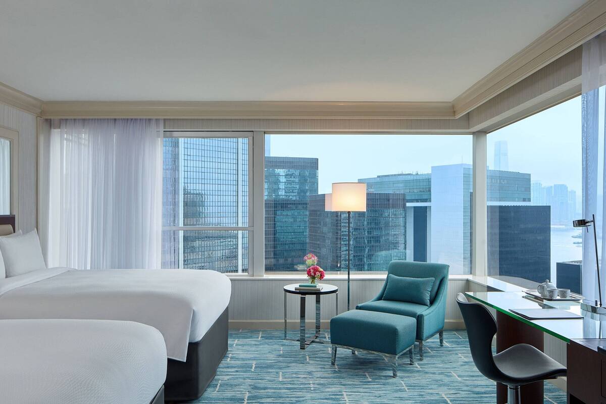 A hotel room with a large bed, a desk, a chair, a small table with a lamp, and a view of the cityscape through large windows.
