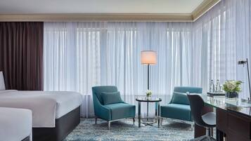 A hotel room with a large bed, two armchairs, a small round table with a lamp, and a desk with a vase of flowers.