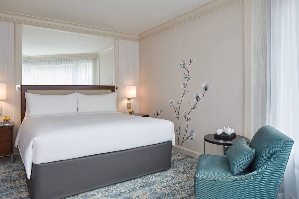 A hotel room with a large bed, a small table with a teapot, a chair, and a wall-mounted floral decoration.
