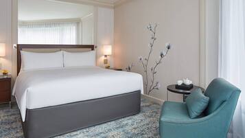 A hotel room with a large bed, a small table with a teapot, a chair, and a wall-mounted floral decoration.
