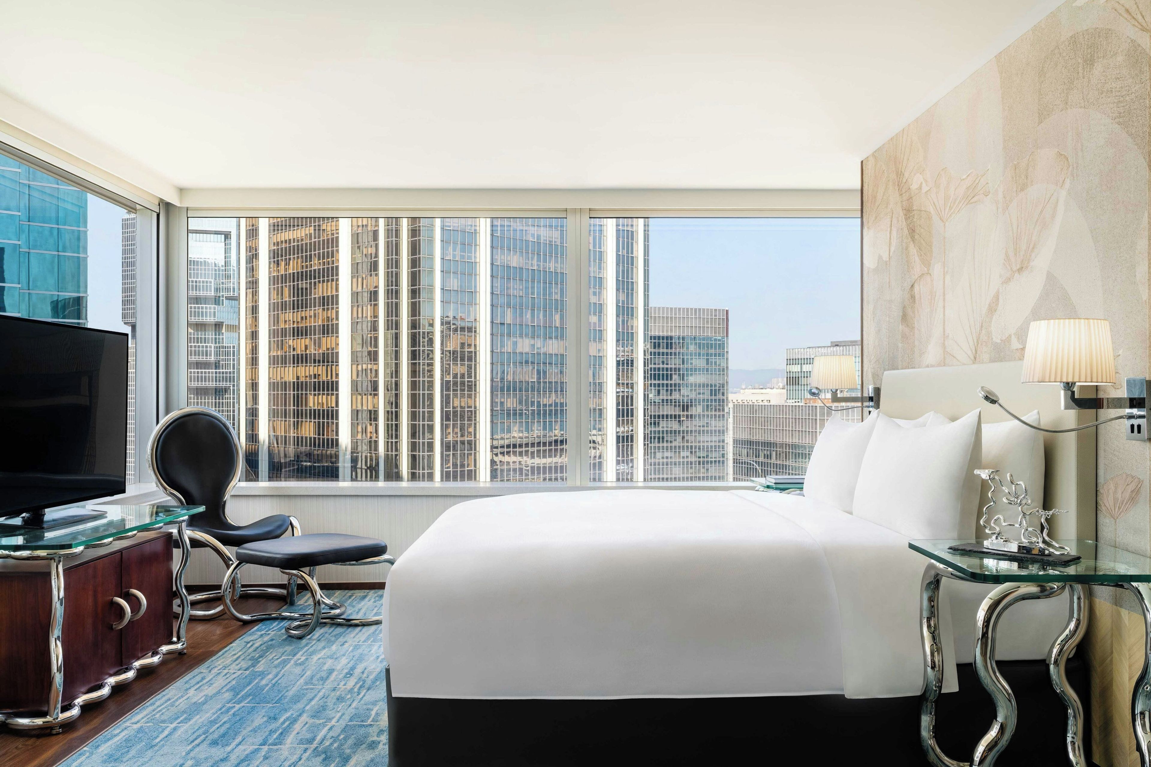 A hotel room with a large bed, a television, a chair, a desk, and a view of city buildings.