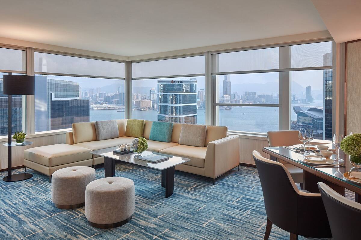 A modern hotel room with a large window offering a city view, a sofa, ottomans, a glass coffee table, and a dining area with chairs and a table set for two.