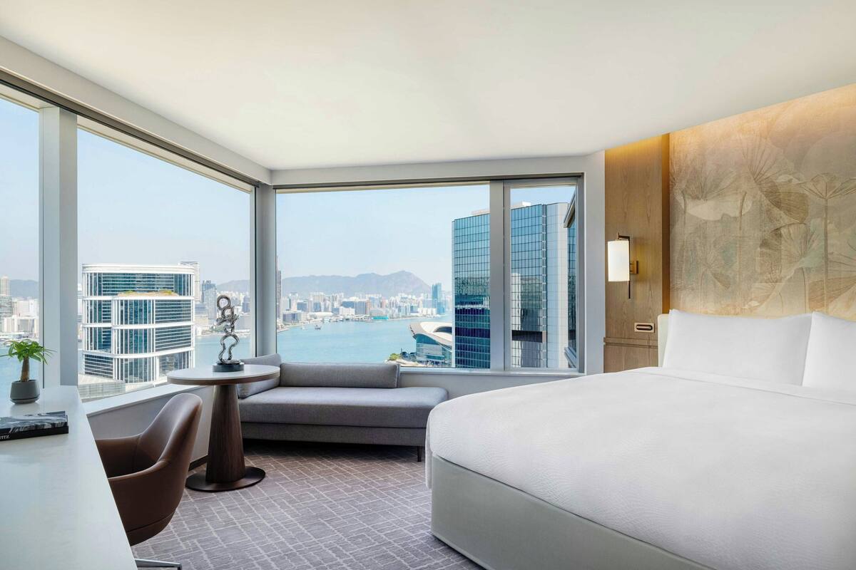 A hotel room with a large window, a bed, a seating area with a table, and a view of a cityscape.
