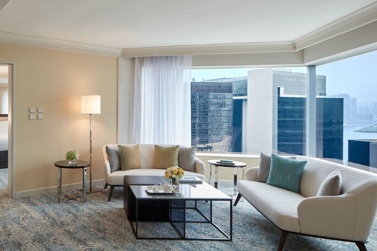 A modern hotel room with a large window offering a city view, a sofa, a coffee table, and a lamp.