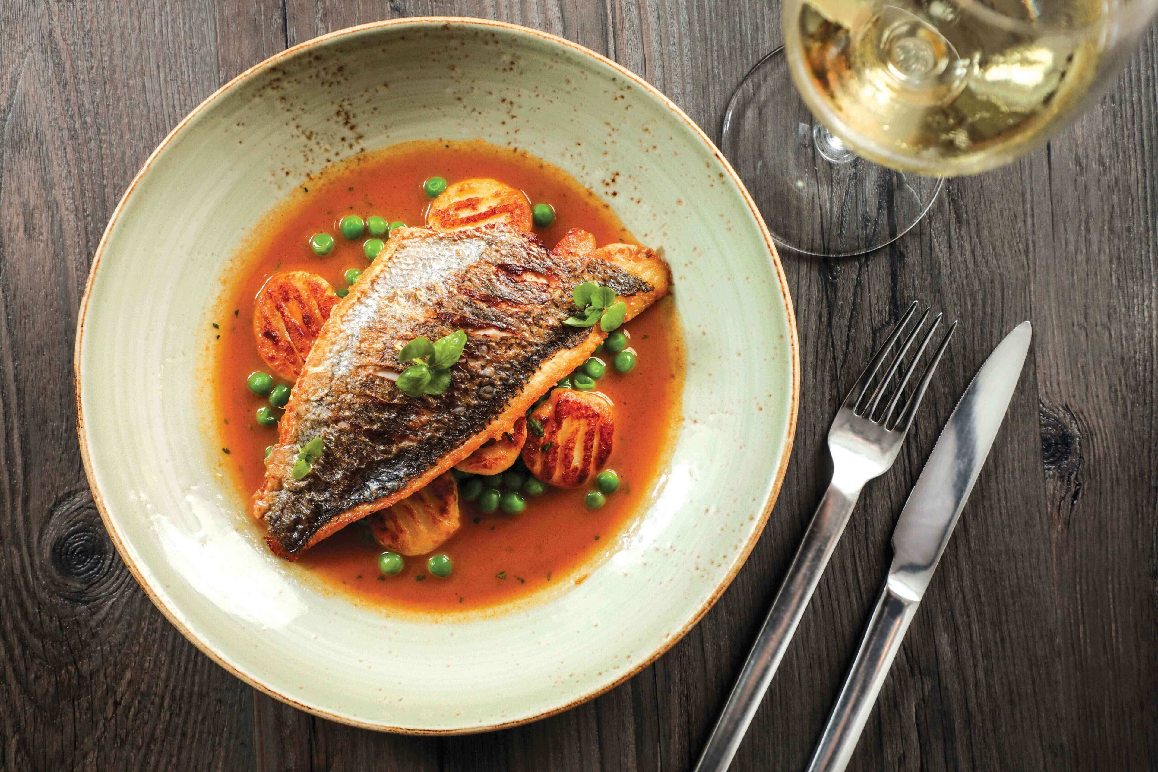 A plate of grilled fish with peas and tomatoes, accompanied by a glass of white wine on a wooden table.