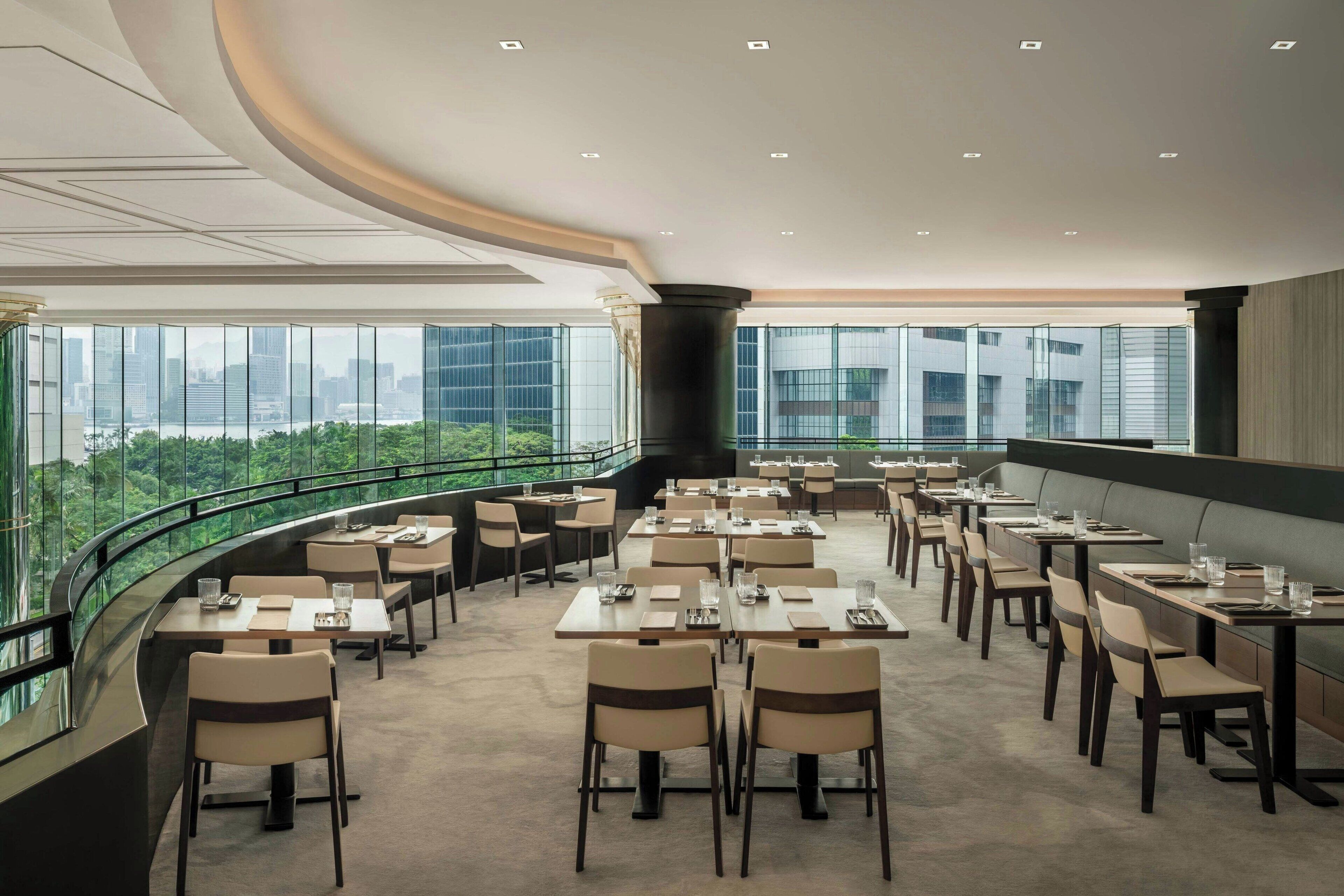 A modern dining area with tables and chairs, large windows offering a city view, and a curved glass railing.