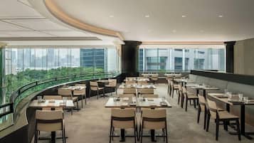 A modern dining area with tables and chairs, large windows offering a city view, and a curved glass railing.