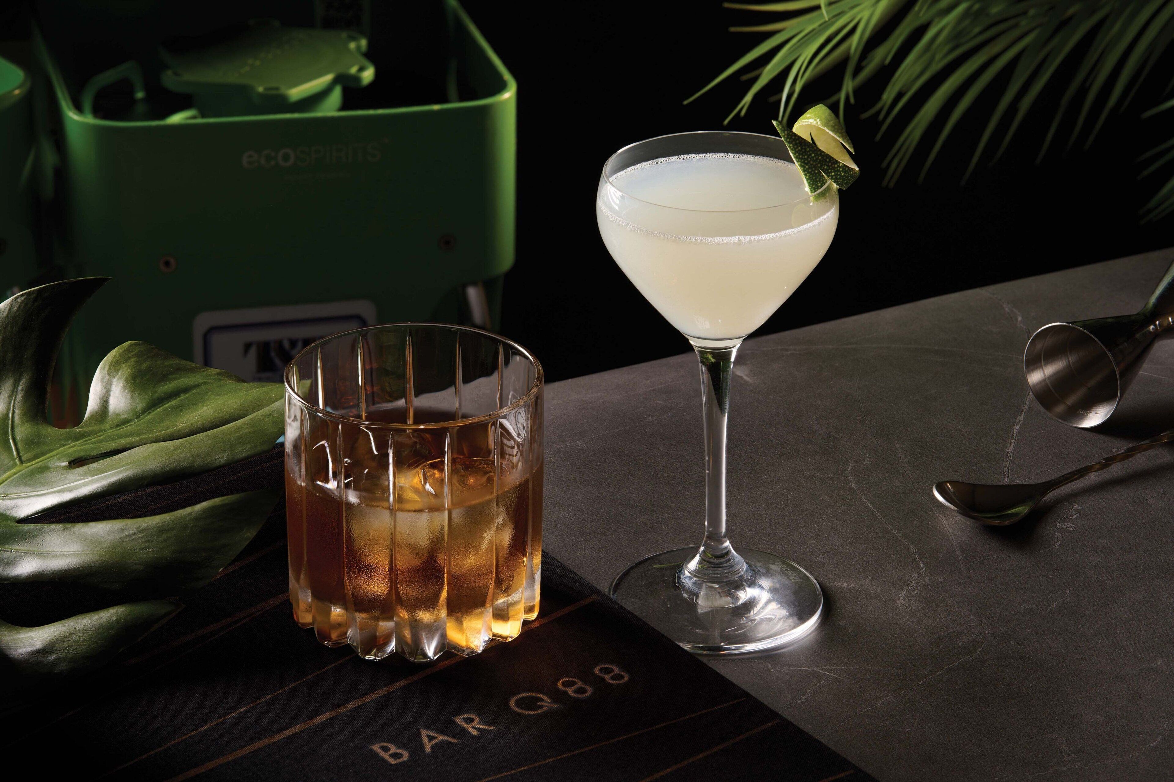 Un bar avec un verre à martini et un verre à whisky sur une surface sombre, un récipient vert portant l'inscription « ecospirit » à l'arrière-plan, et un menu avec le texte « BAR Q88 ».