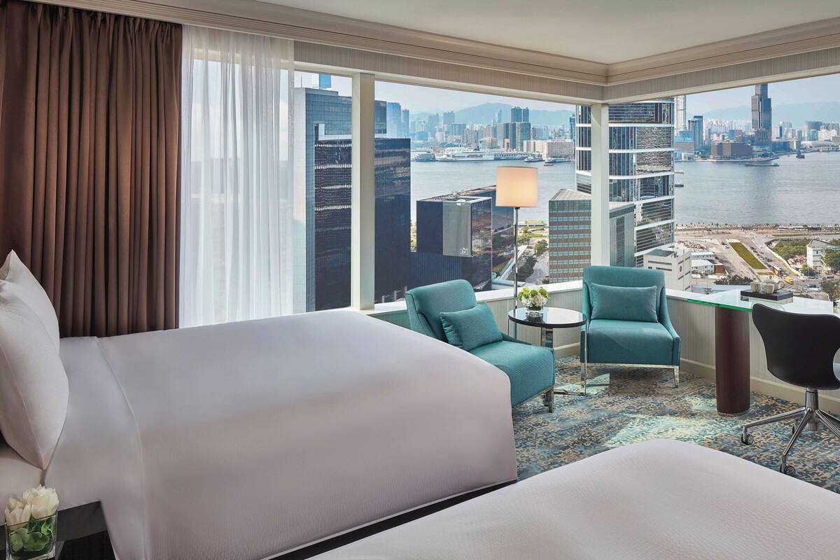 A hotel room with a large bed, a desk, two chairs, and a view of a cityscape and river.