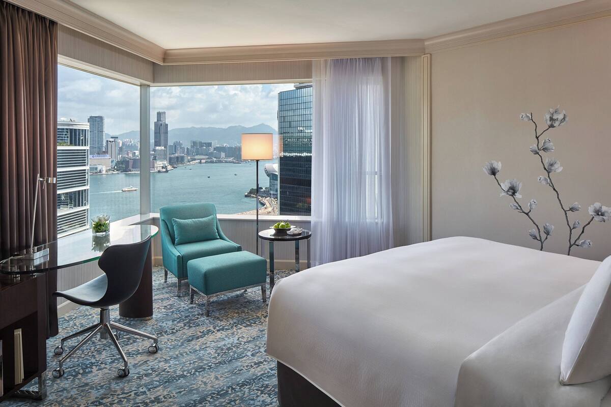 A hotel room with a large bed, a desk with a chair, a small table with a lamp, and a view of a cityscape and water.