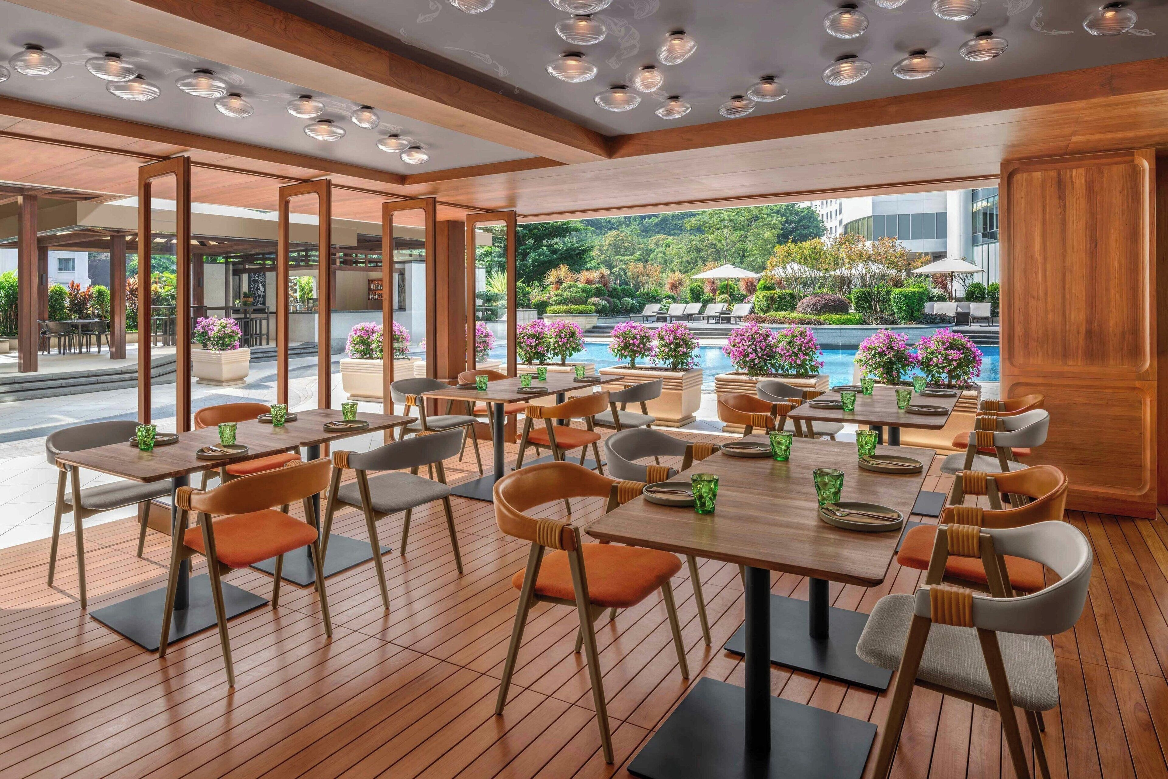 A modern restaurant with wooden flooring, round tables, and orange chairs. It features large windows with a view of a pool and greenery.