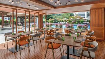 A modern restaurant with wooden flooring, round tables, and orange chairs. It features large windows with a view of a pool and greenery.