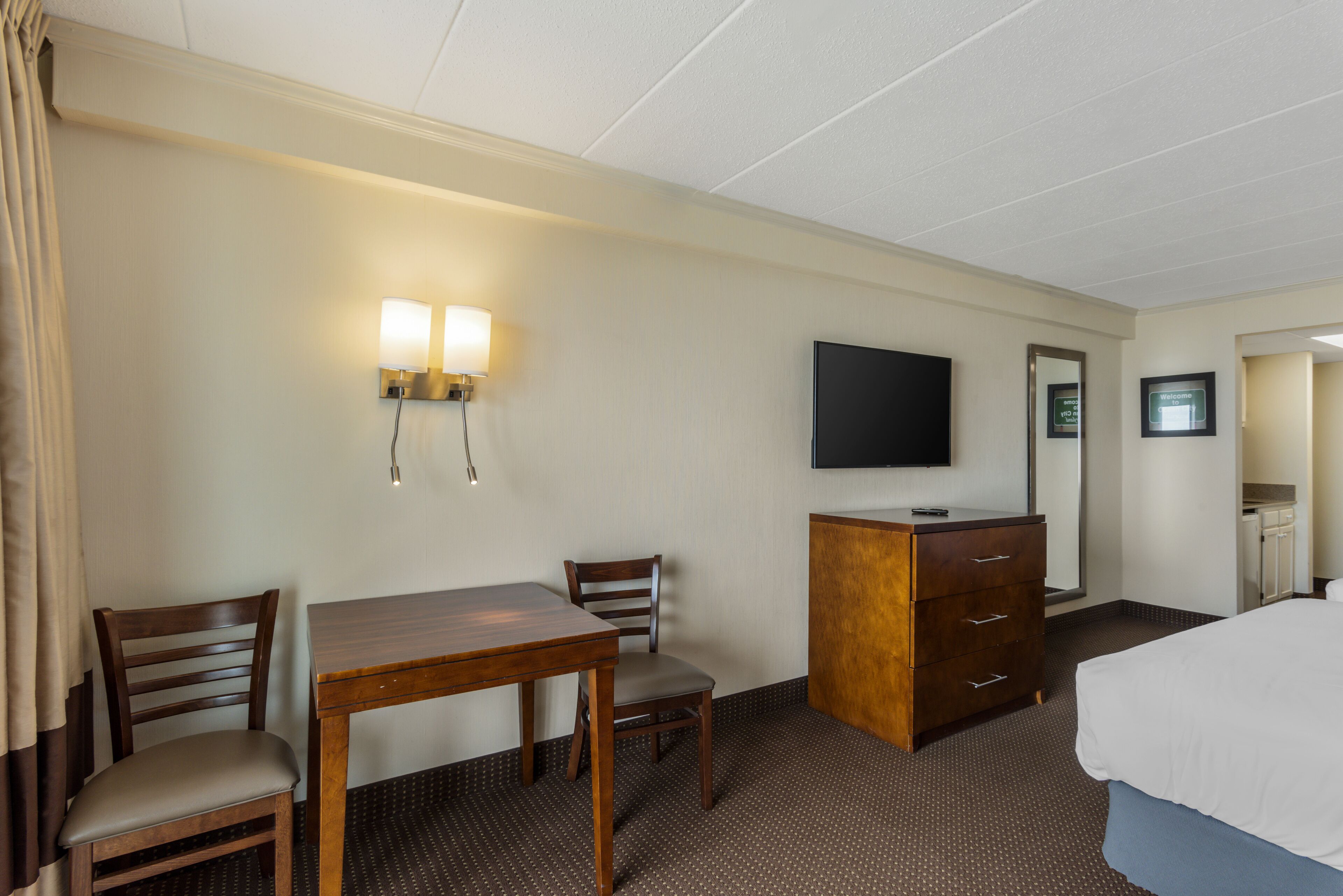 A hotel room with a bed, a desk with two chairs, a dresser, a flat-screen TV mounted on the wall, and a wall-mounted light fixture.