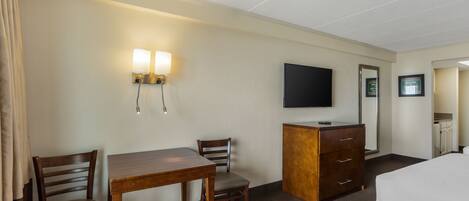 A hotel room with a bed, a desk with two chairs, a dresser, a flat-screen TV mounted on the wall, and a wall-mounted light fixture.
