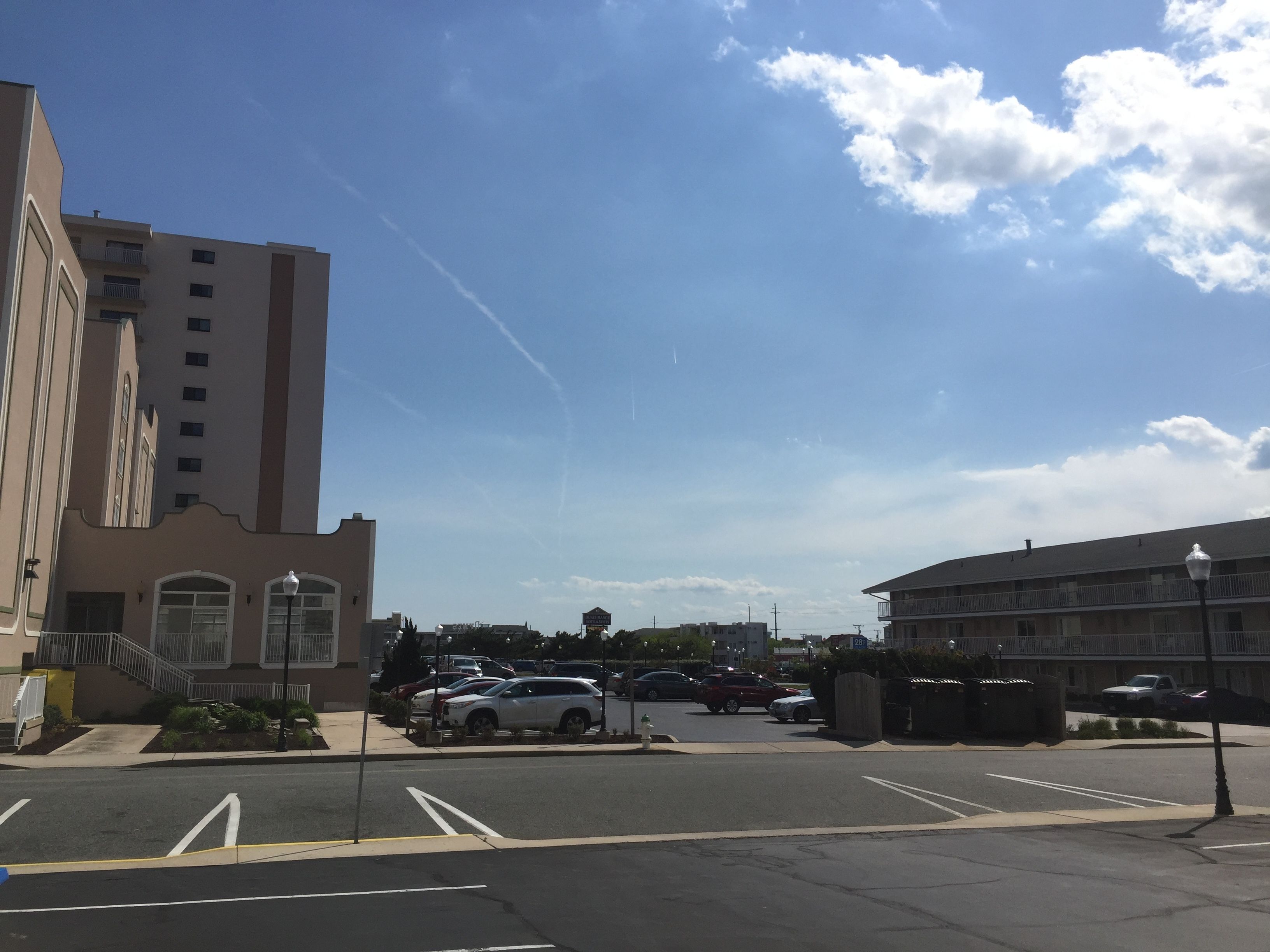 Um estacionamento com vários carros, um edifício com vários andares e um céu claro com algumas nuvens.