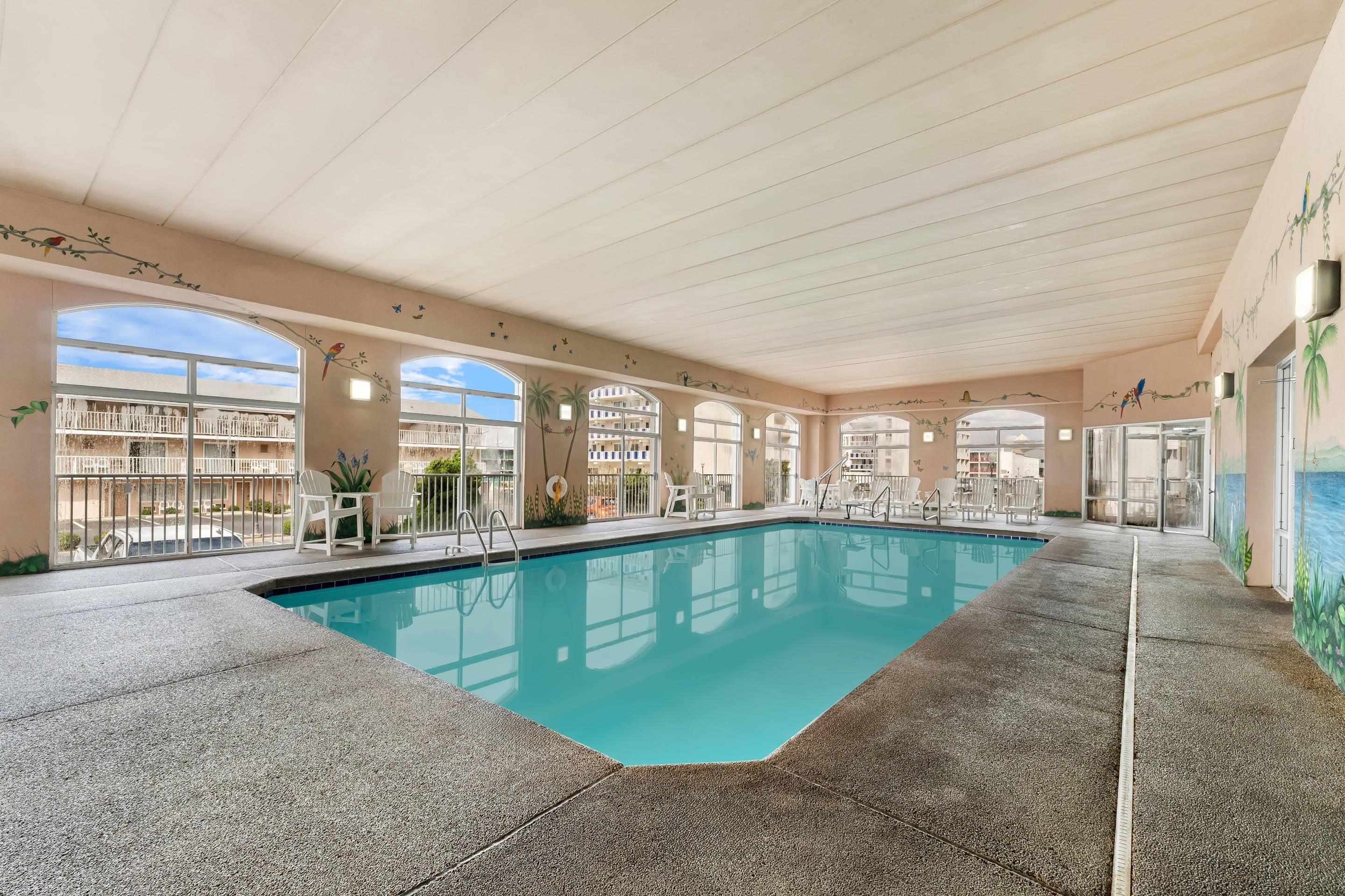An indoor swimming pool with clear blue water, surrounded by white chairs and tables, and large windows offering views of the outside.