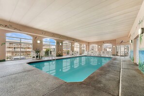 An indoor swimming pool with clear blue water, surrounded by white chairs and tables, and large windows offering views of the outside. - Quality Inn Ocean City Beachfront (Ocean City)