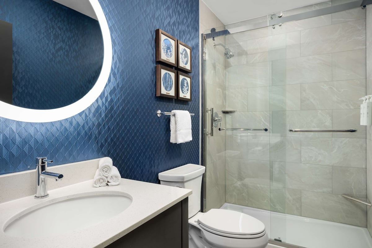 Un baño moderno con una pared de azulejos azules, un espejo redondo, un lavabo blanco y una ducha con mampara de vidrio.