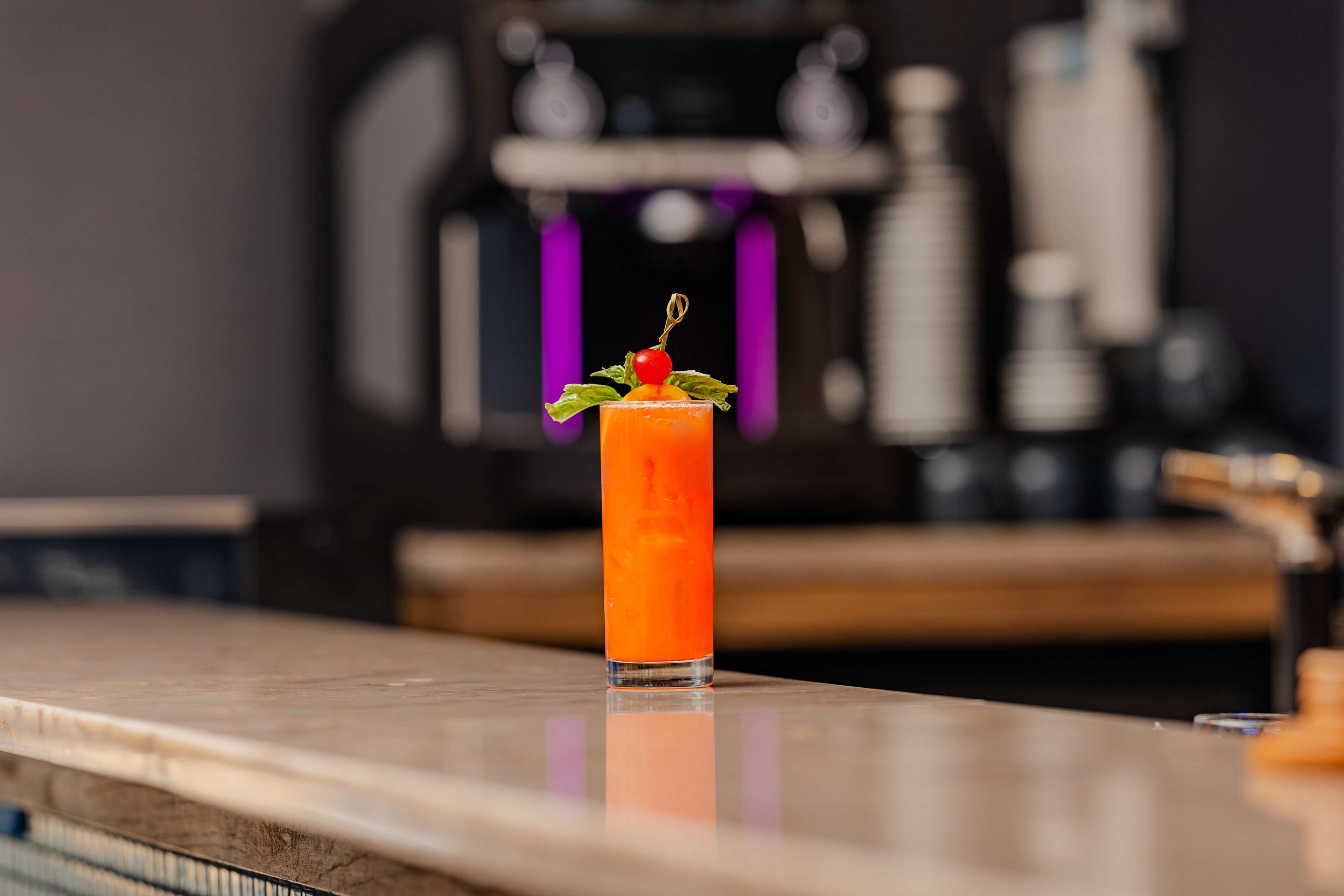 A tall glass of orange-colored drink with a cherry and mint garnish on a reflective surface.