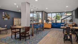 A modern dining area with wooden tables and chairs, a blue patterned rug, and large windows offering a view of the outside.