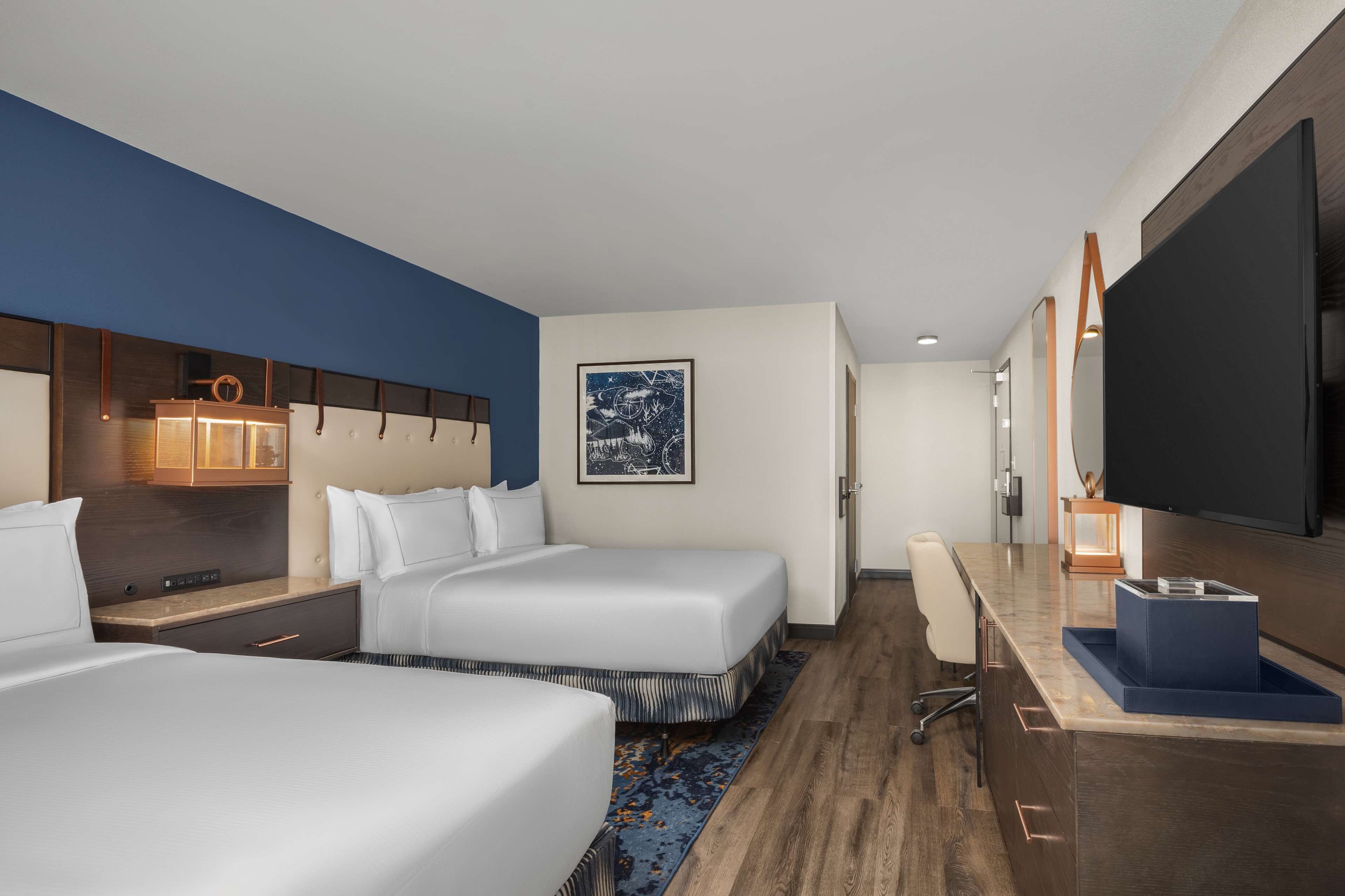 A hotel room with two beds, a desk, a flat-screen TV, and a large window.