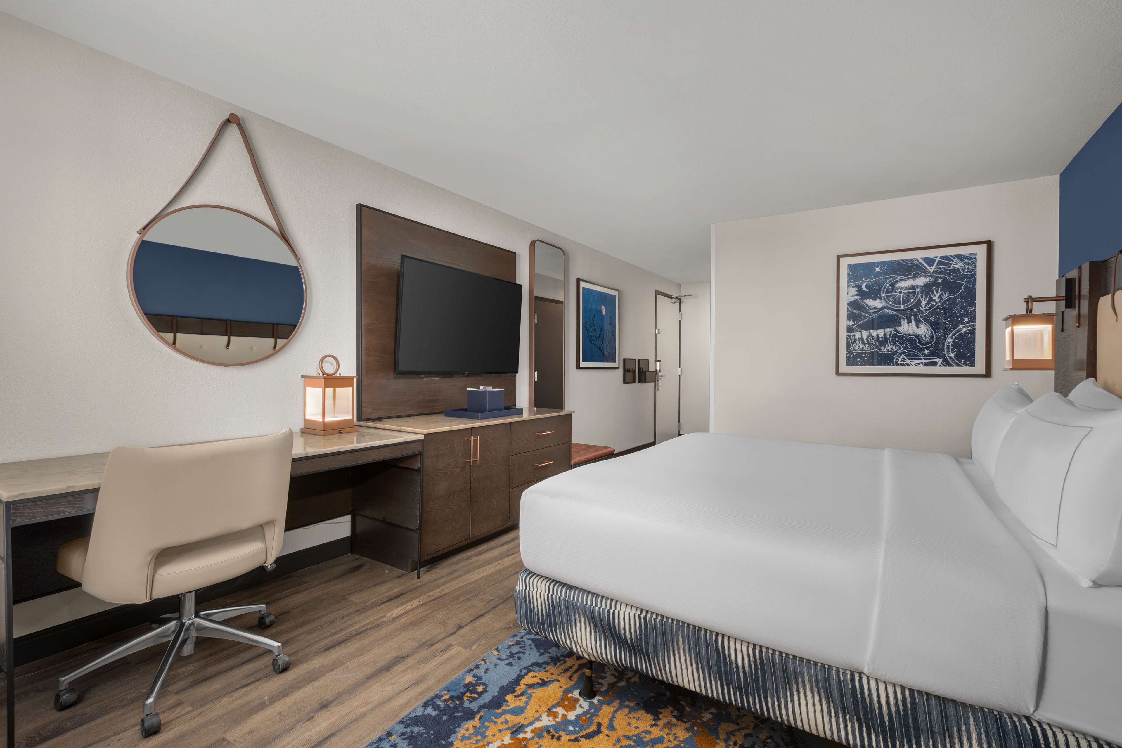 A modern hotel room with a large bed, a desk with a chair, a television, and a mirror.