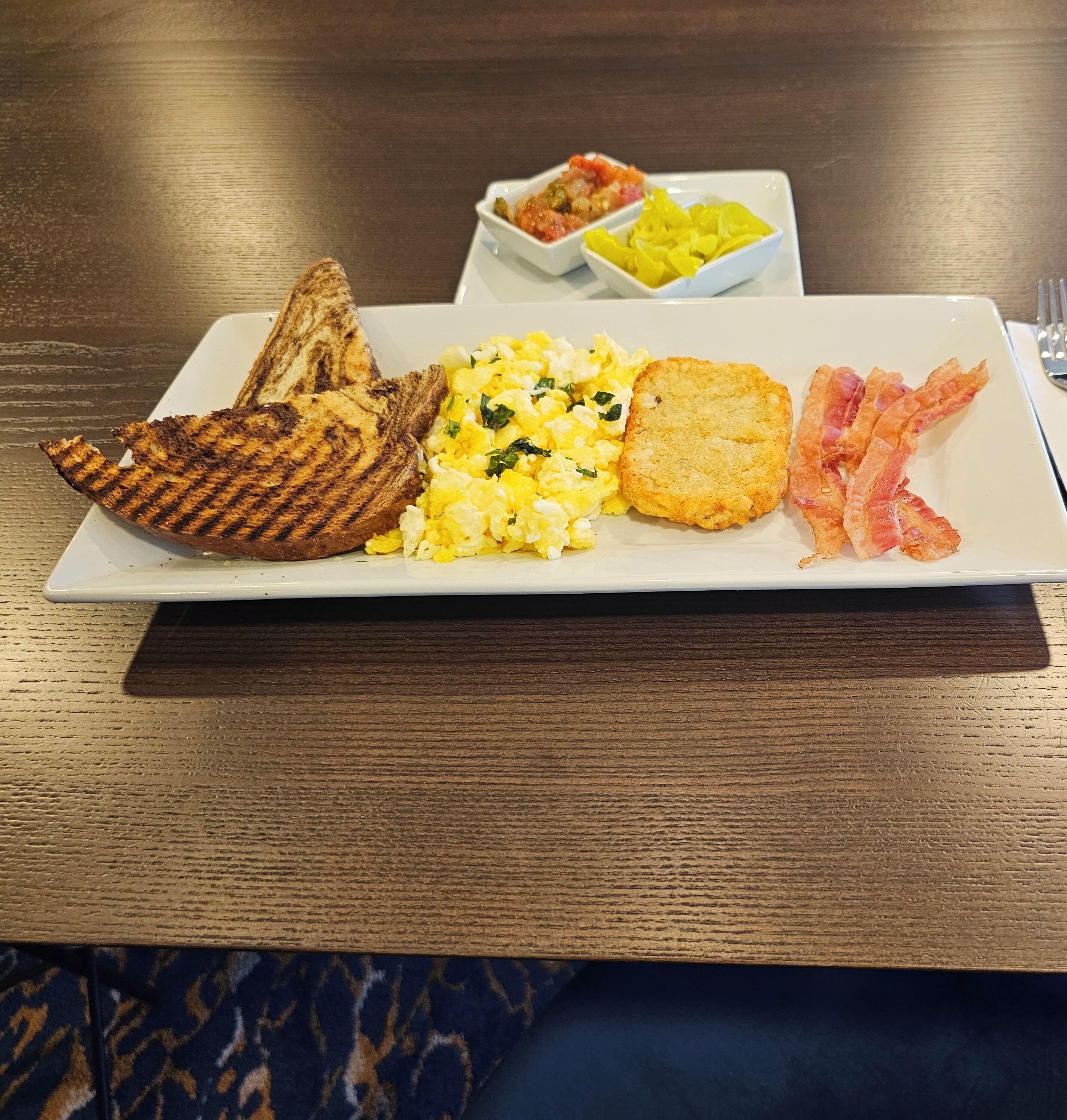 A breakfast plate with scrambled eggs, toast, bacon, and a side of fruit.