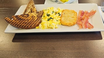 A breakfast plate with scrambled eggs, toast, bacon, and a side of fruit.
