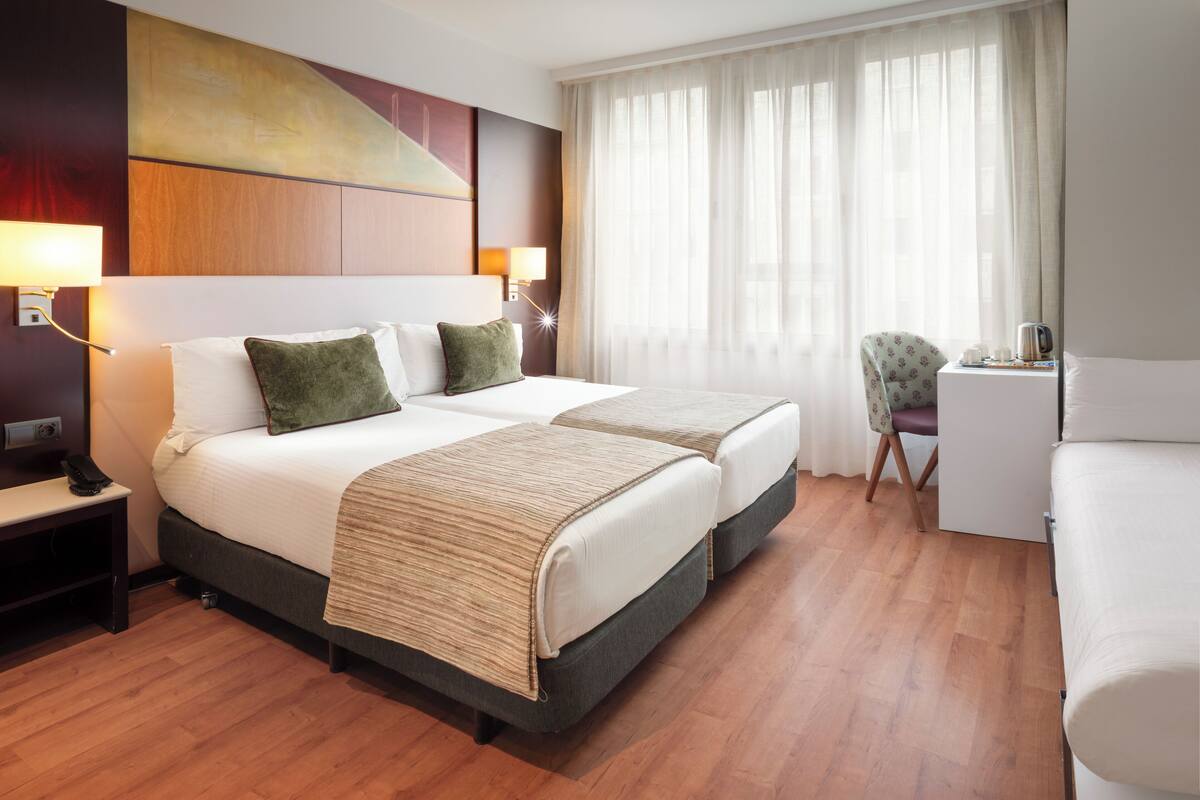 Habitación de hotel con dos camas, un escritorio, una silla y un ventanal con cortinas.