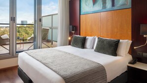 A modern hotel room with a large bed, a balcony with a table and chairs, and a city view.