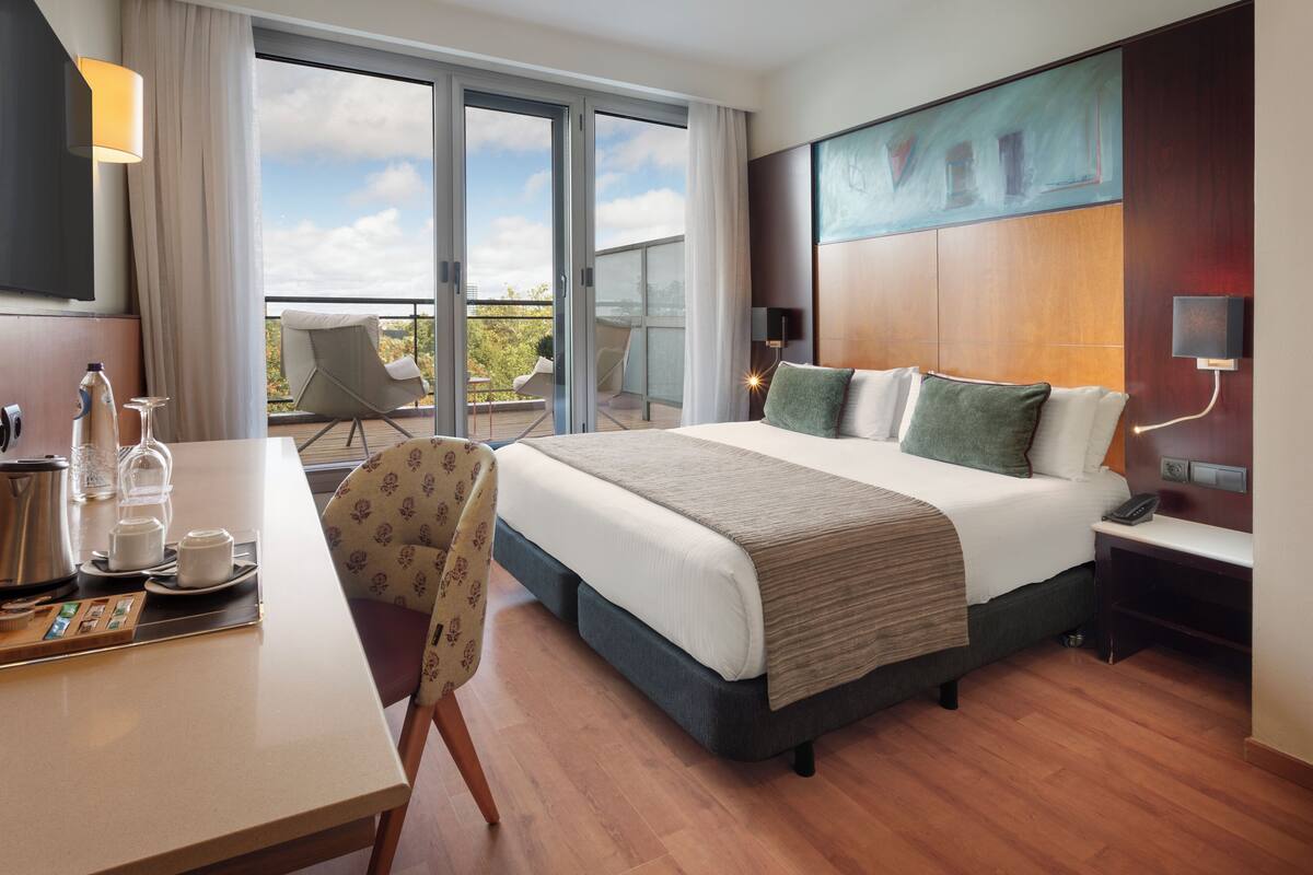 Habitación de hotel con cama, escritorio con utensilios para preparar café, una silla y balcón con un sillón.