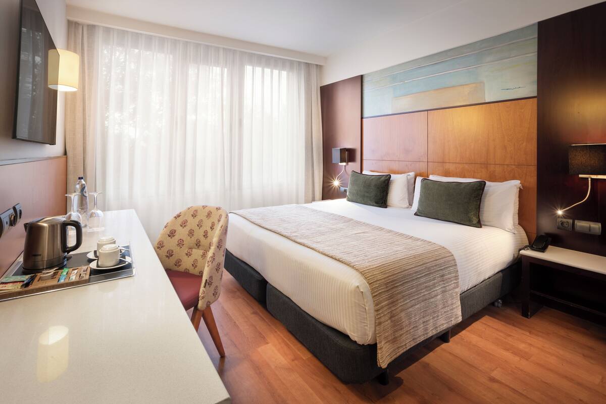 Una habitación de hotel con cama, mesita de noche, lámpara y escritorio con tetera y tazas.