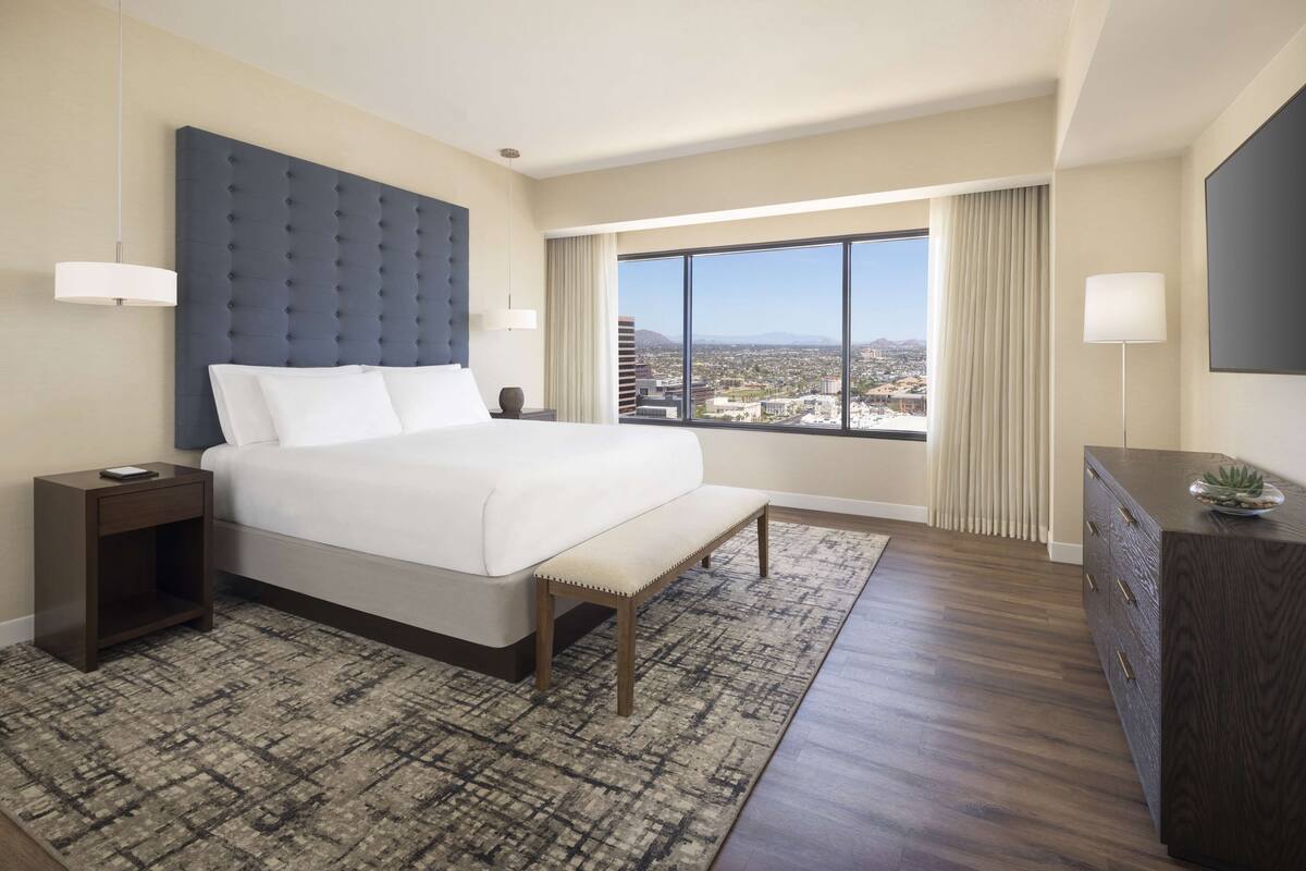A hotel room with a large bed, a view of the city, a desk, and a TV.