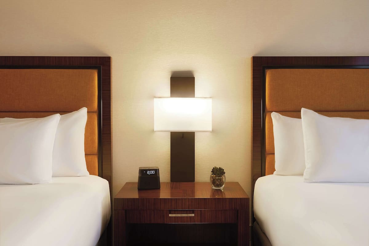 A hotel room with two beds, a nightstand with a clock, a lamp, and a small plant.