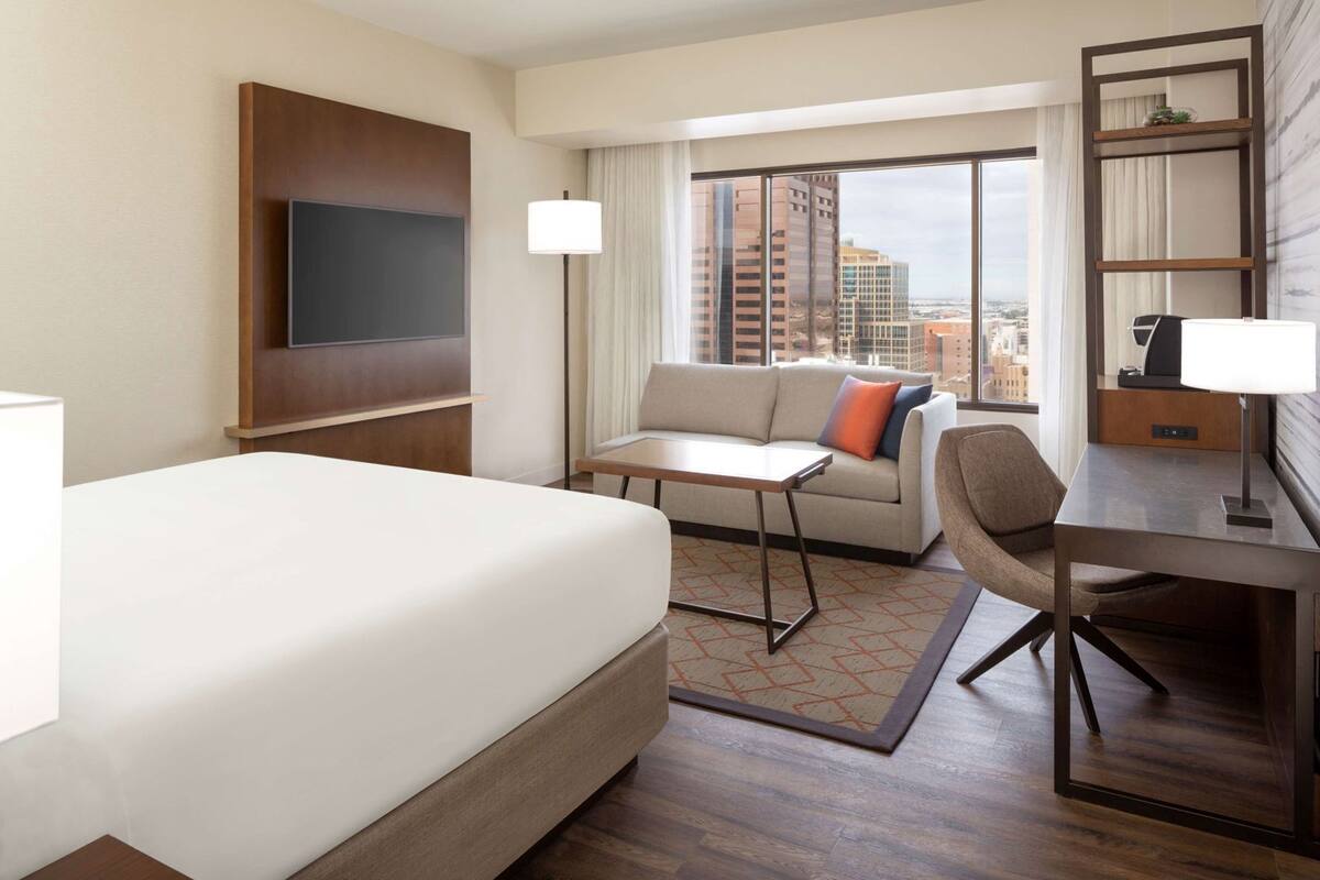 A hotel room with a bed, a sofa, a desk, a TV, and a window with a city view.