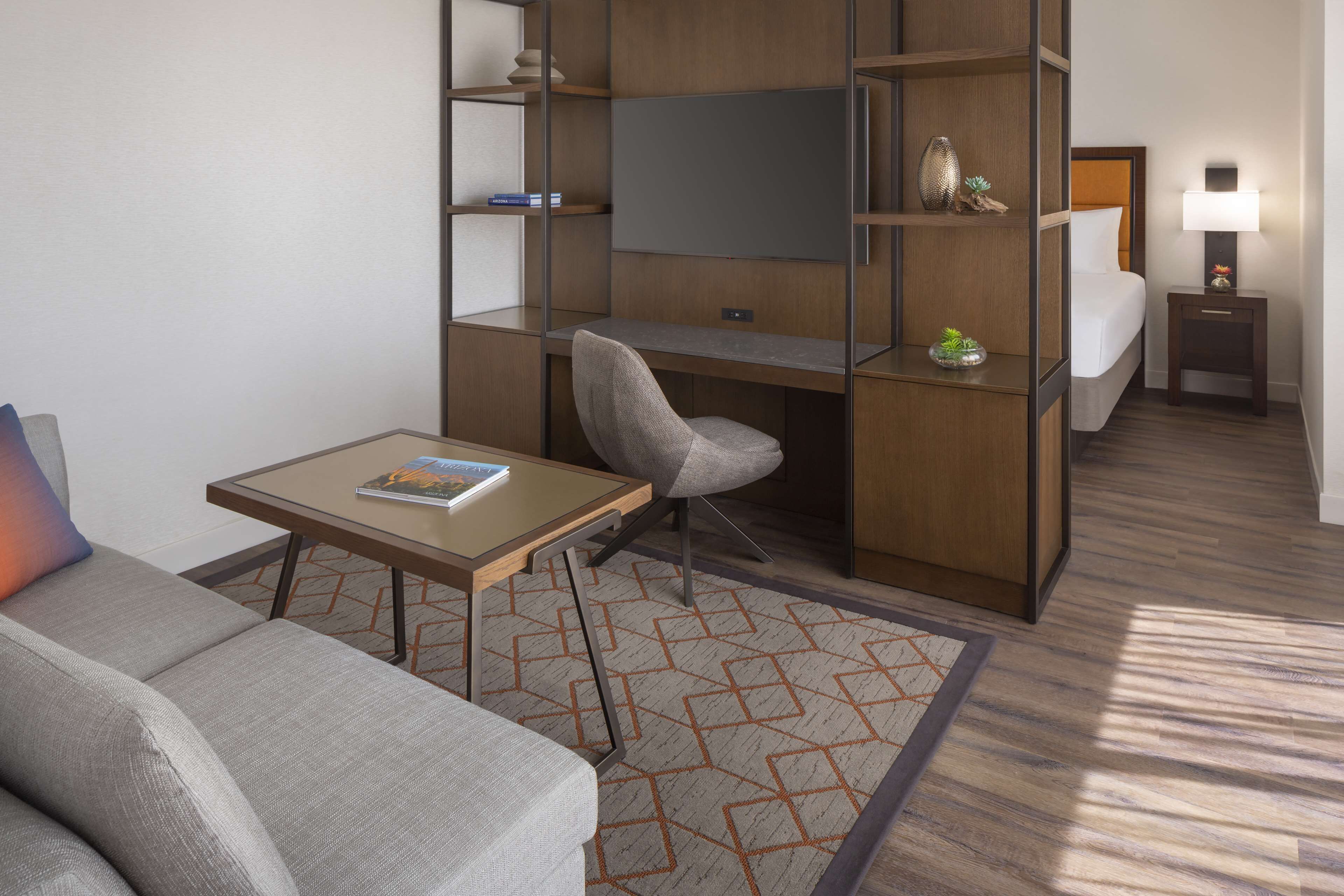 A compact hotel room with a sofa, a small table, a desk with a chair, a bed, a nightstand with a lamp, and built-in shelving.