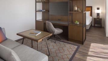 Habitación de hotel compacta con sofá, una pequeña mesa, escritorio con silla, cama, mesita de noche con lámpara y estanterías empotradas.