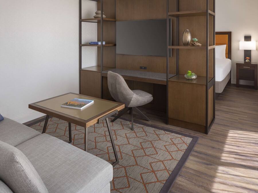 A compact hotel room with a sofa, a small table, a desk with a chair, a bed, a nightstand with a lamp, and built-in shelving.