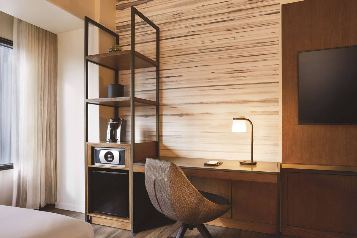 A hotel room with a bed, a desk with a lamp, a chair, a TV, and a minibar.