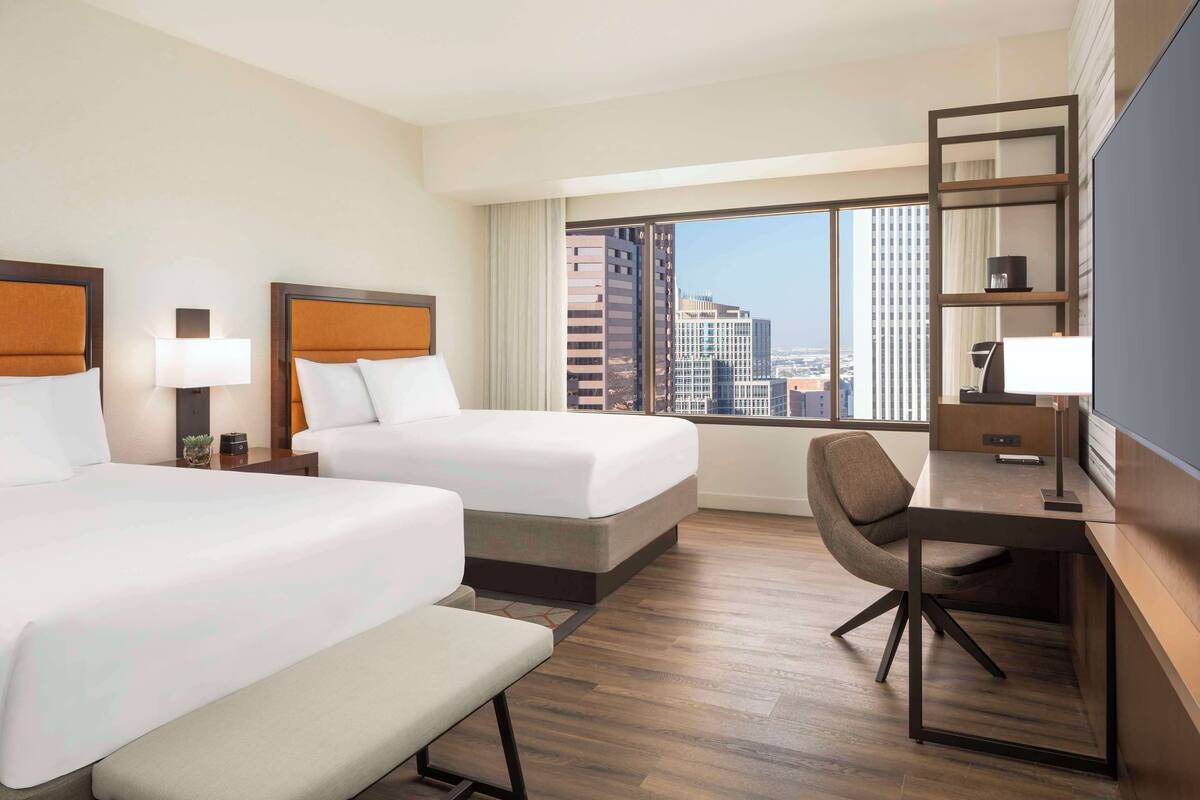 A hotel room with two beds, a desk, a chair, a TV, and a large window with a city view.