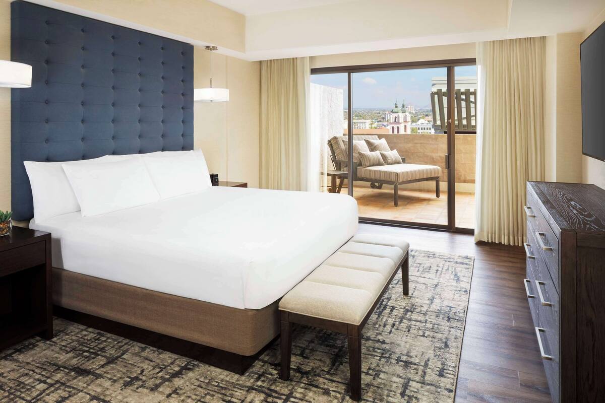 A hotel room with a large bed, a bench, a dresser, a chair, and a balcony with a view.