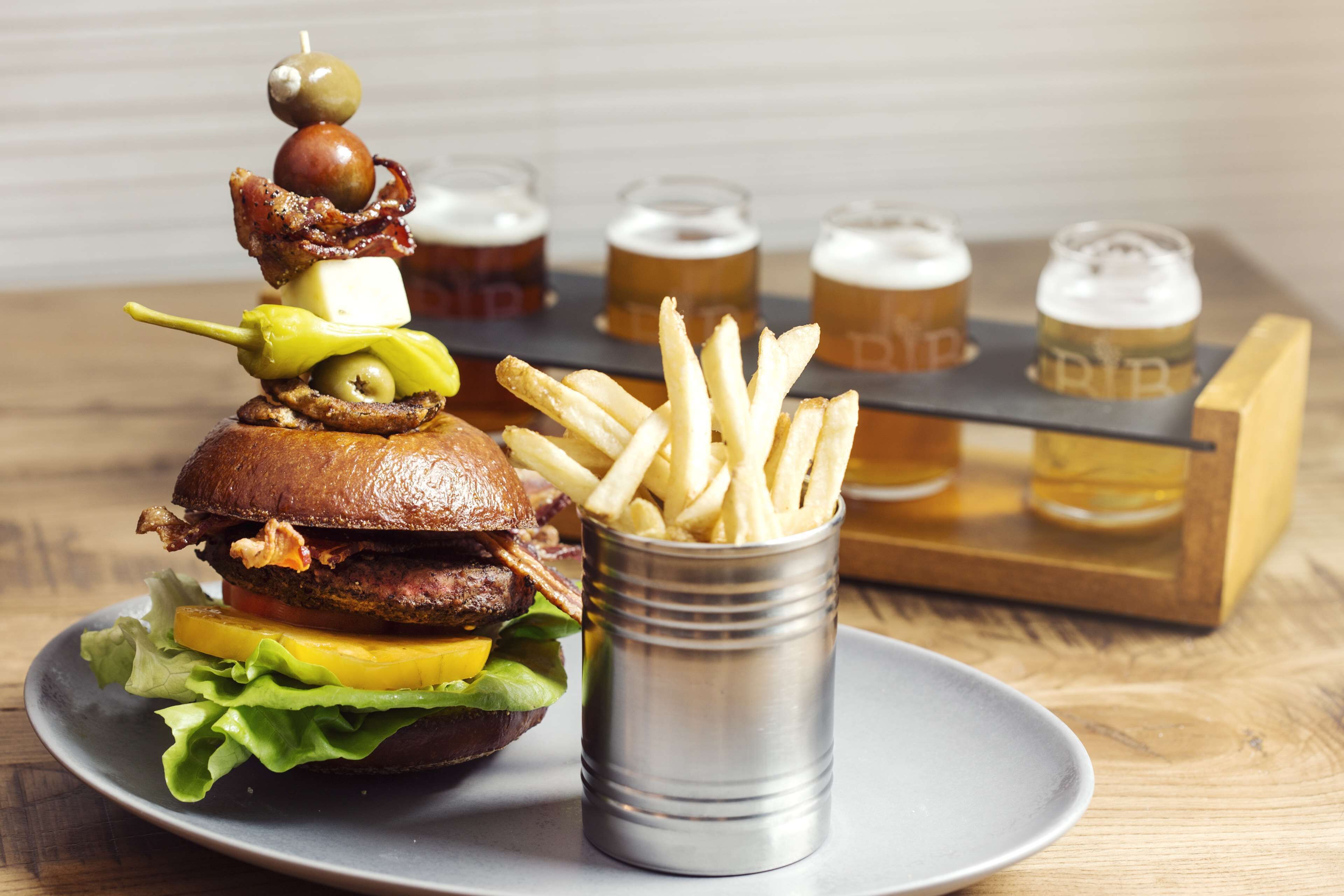 En hamburgare med bacon, ost och inlagd gurka, serverad på ett bröd, tillsammans med en portion pommes frites och fyra ölglas på ett bricka.