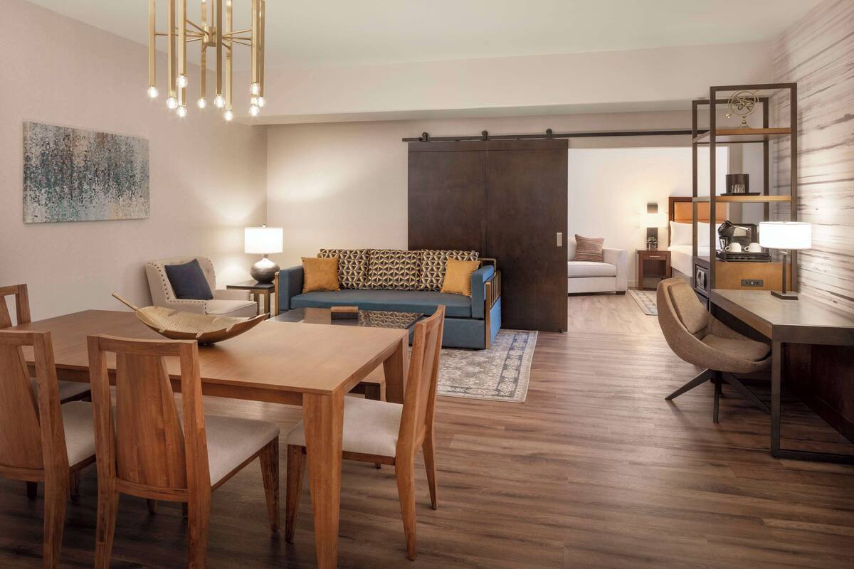 A modern living room with a sofa, dining table, and chairs. There is a sliding door closet and a small desk with a lamp.