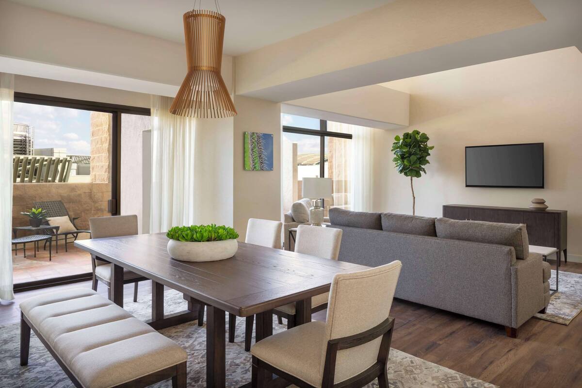A spacious living area with a wooden dining table, cushioned chairs, a grey sofa, and a large window offering a view of the outdoors.