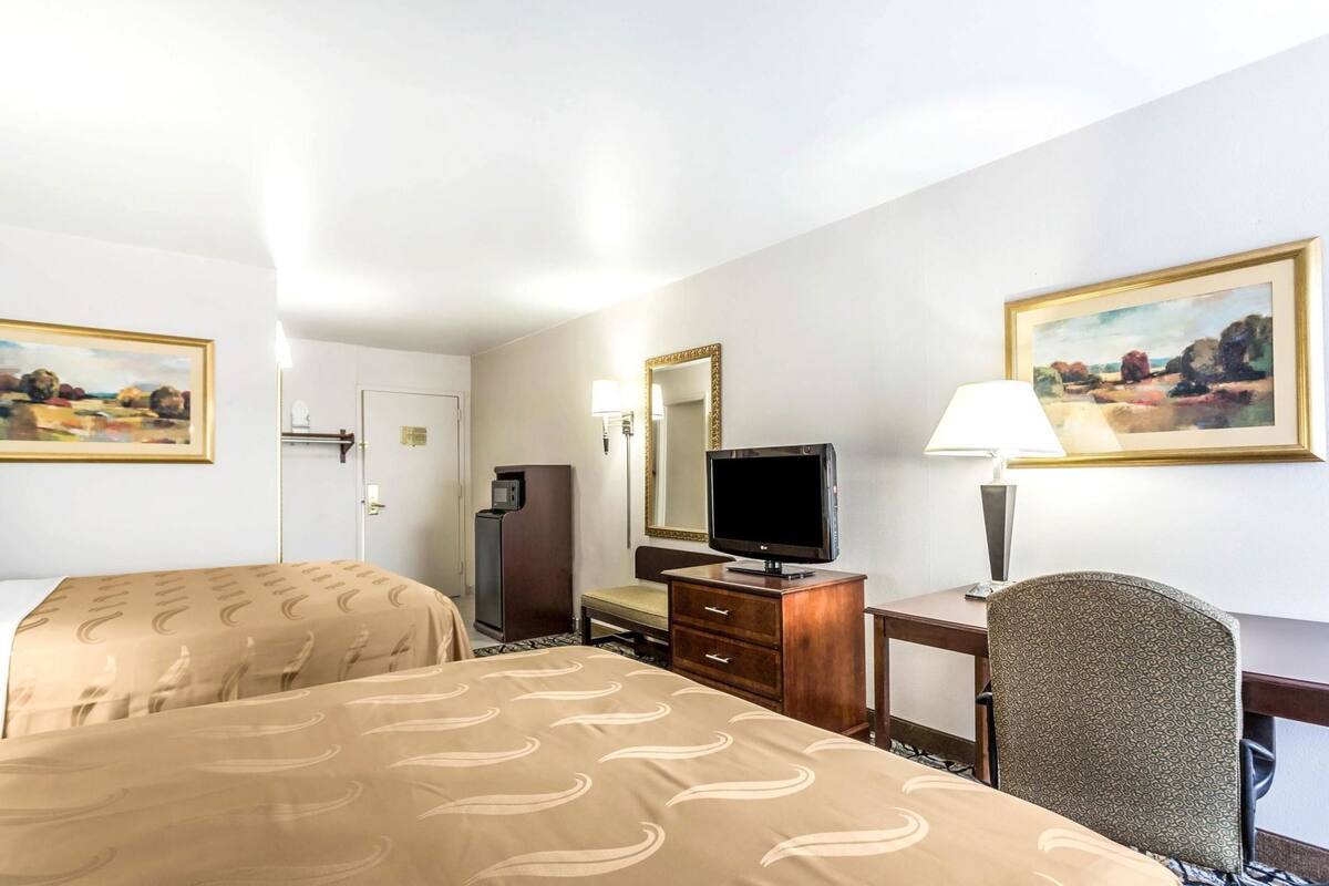 A hotel room with two beds, a desk, a television, a mirror, and artwork on the walls.