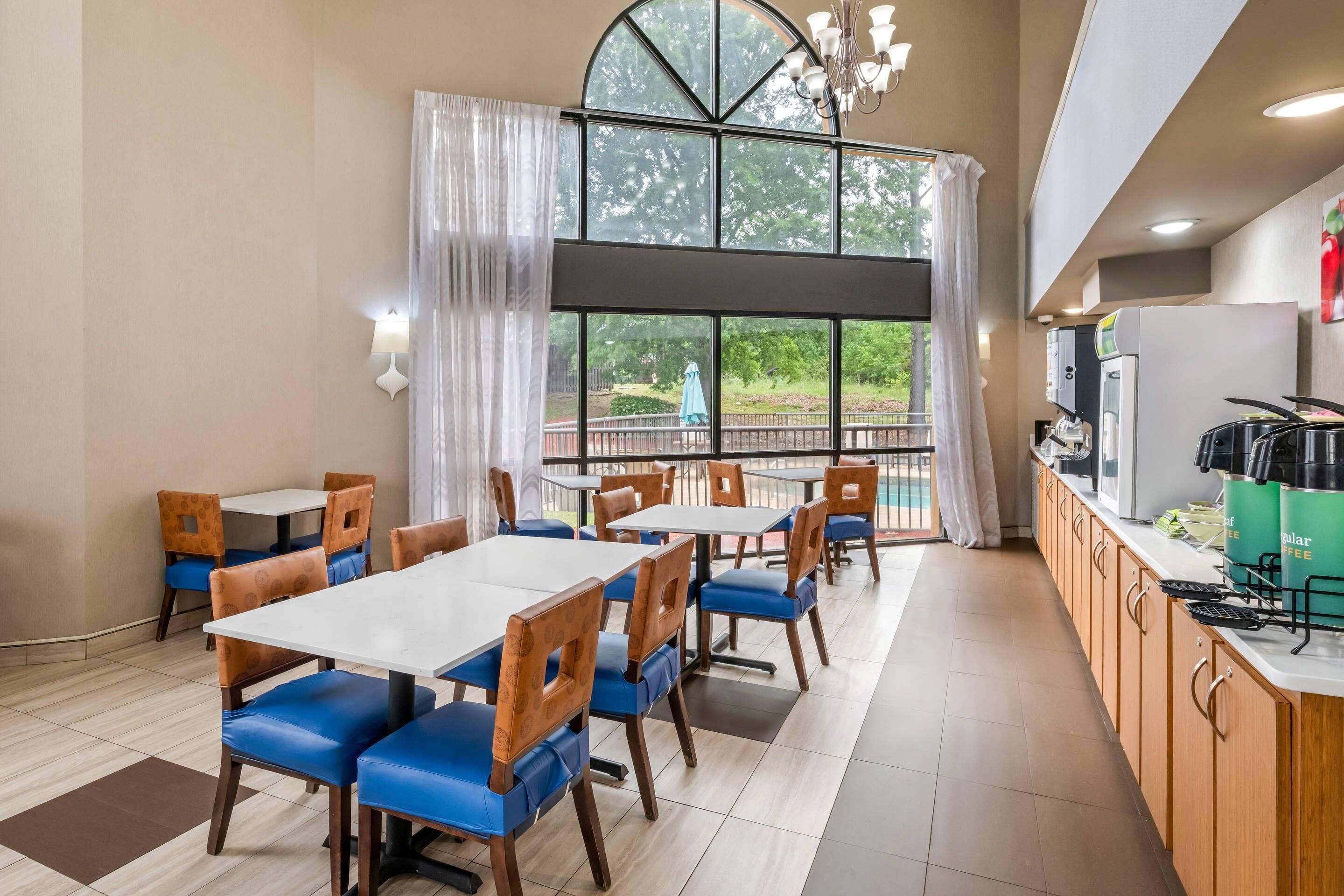 A restaurant with tables and chairs, a kitchen area, and large windows with a view of greenery.
