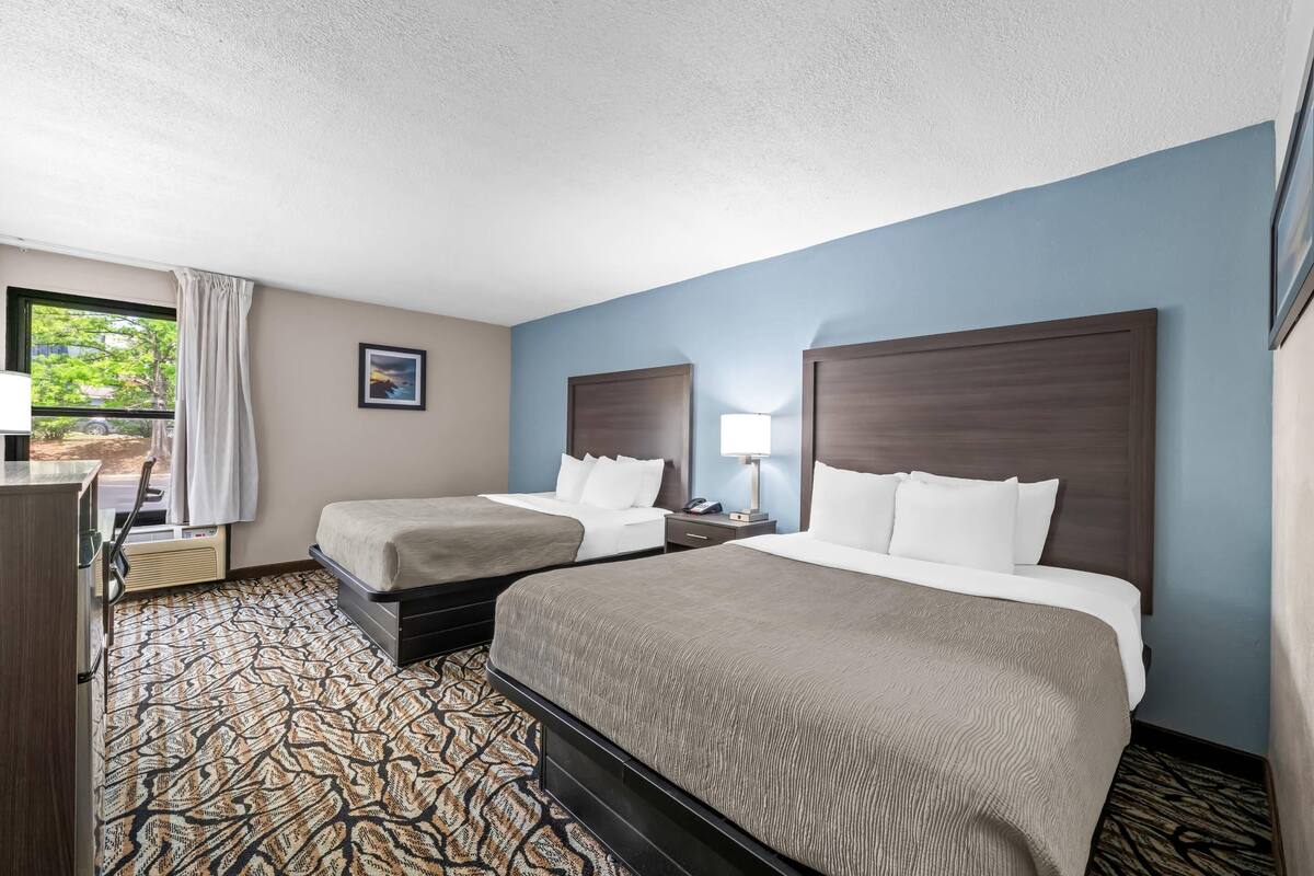 A hotel room with two beds, a desk, a chair, a window with curtains, and a patterned carpet.