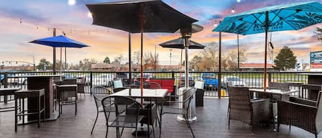 An outdoor patio with tables and chairs, umbrellas, and string lights against a sunset sky.